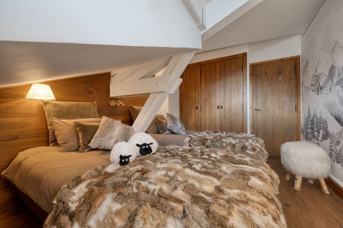 A cozy attic bedroom features a wooden headboard, a fur throw on the bed, decorative sheep pillows, and a mountain mural, creating a warm chalet ambiance.