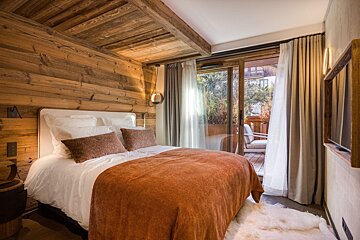 A cozy, rustic bedroom with wooden walls and ceiling, a large bed with an orange blanket, and a sliding glass door opening to an outdoor patio.