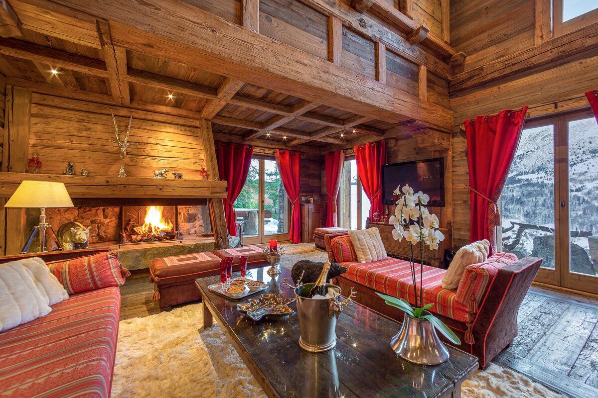 Luxurious wooden chalet living room with a cozy fireplace, red accents, and stunning views of a snowy mountain landscape through large windows.