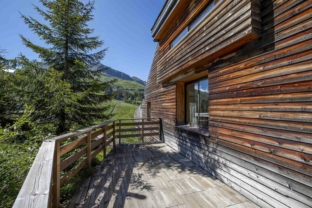 A modern, wood-paneled living space with a yellow sofa and wooden dining table, featuring large windows offering expansive views of a lush green landscape.