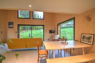 A bright, modern wooden cabin interior featuring a yellow sofa, dining table, and large windows looking out onto a green lawn.