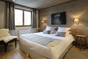 Chalet bedroom with rustic wood walls/ceiling, a plush white bed, and a striking portrait painting. Warm lighting and a sheepskin chair add coziness.