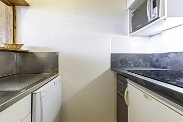 A compact kitchen with white walls, stainless steel sink, dishwasher, dark counter with built-in hob, under-counter fridge, and an overhead microwave.