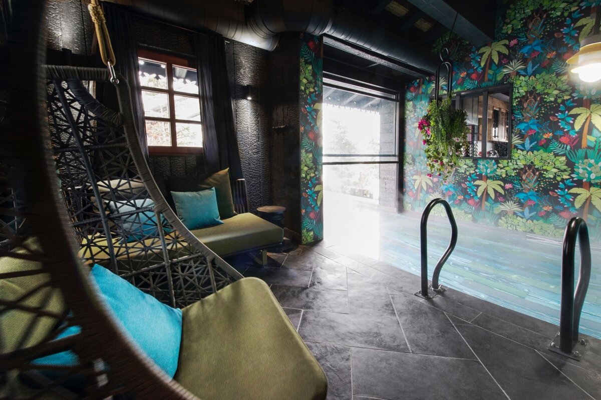A tropical-themed indoor pool area with dark walls, vibrant jungle wallpaper, a hanging chair, and a chaise lounge with teal pillows.