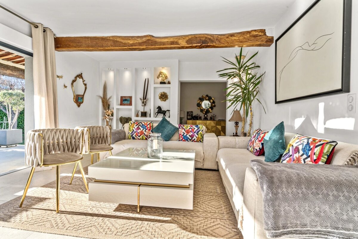 A bright, stylish living room featuring a large beige sectional, white coffee table, and decorative shelves. A rustic wooden beam and colorful pillows add character.