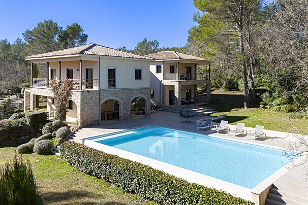 A luxurious two-story villa with a large, clear blue swimming pool, lounge chairs, and surrounding lush greenery.