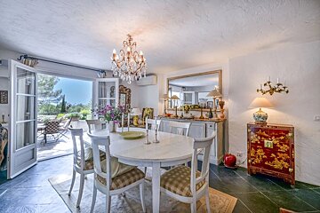 An elegant dining room featuring a white wooden table, a crystal chandelier, and open French doors leading to a sunny terrace.