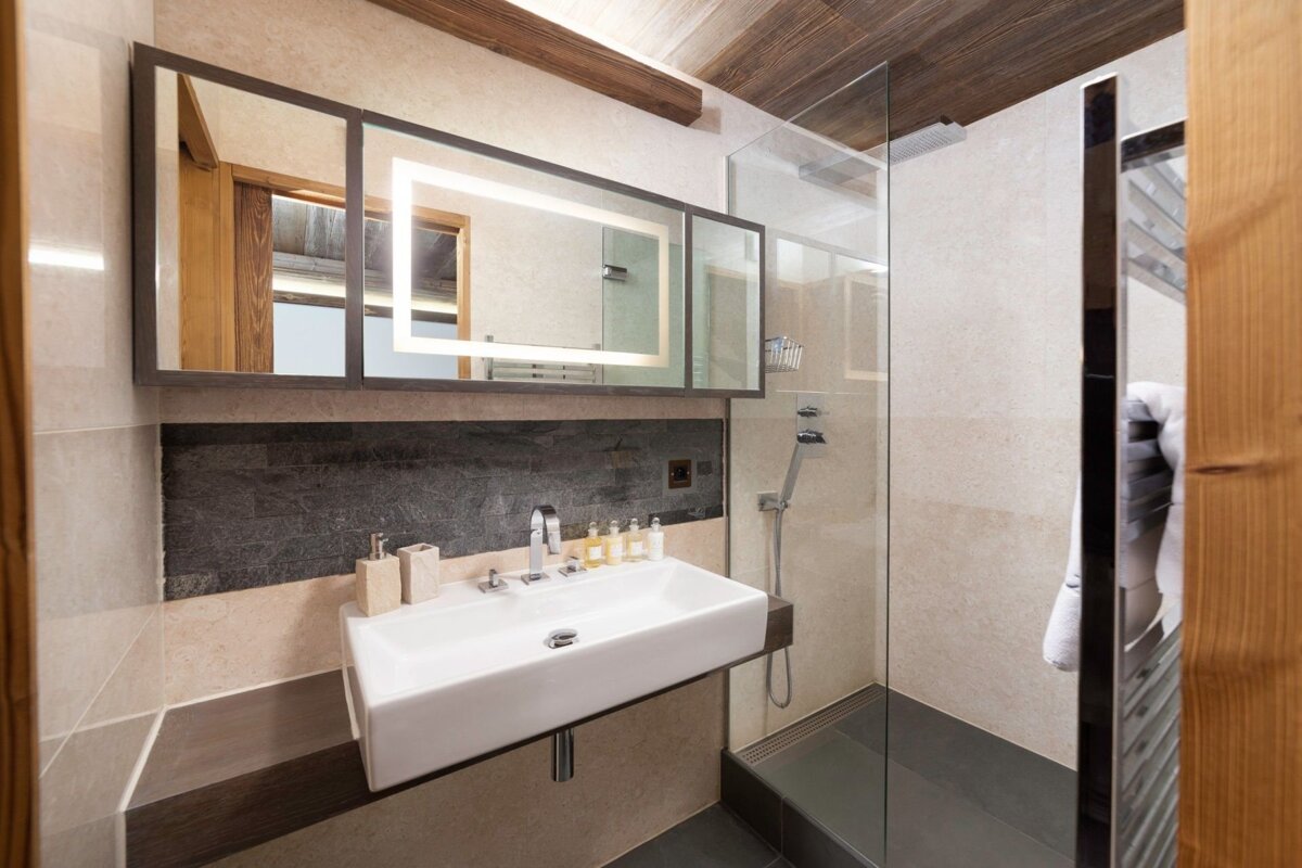 A modern bathroom featuring a rectangular sink, illuminated mirror, glass shower, and tiled walls with wooden ceiling details.