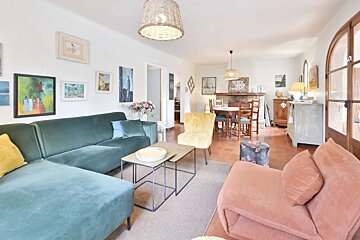 A vibrant living/dining room with a teal sectional, peach chaise, and yellow chair. Art fills the walls, and woven light fixtures hang above.
