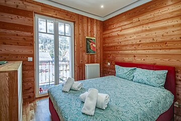 Cozy wood-paneled bedroom with a red bed, teal patterned bedding, and a balcony overlooking a mountain town. Fresh towels are ready.
