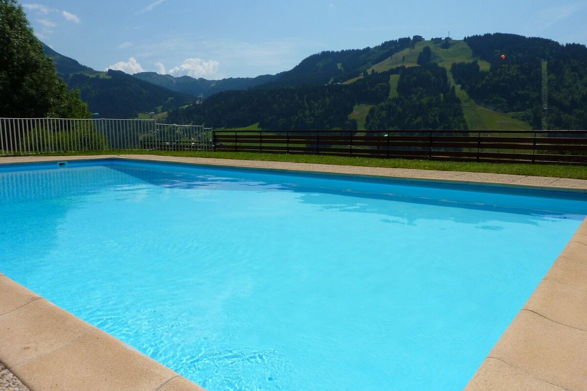 A bright blue swimming pool overlooks lush green mountains with ski slopes under a sunny sky, offering a picturesque summer view.