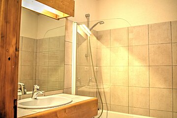 A warm-toned bathroom featuring a sink, mirror, and tiled shower/bathtub combo with a glass screen. Wooden elements add a rustic touch.