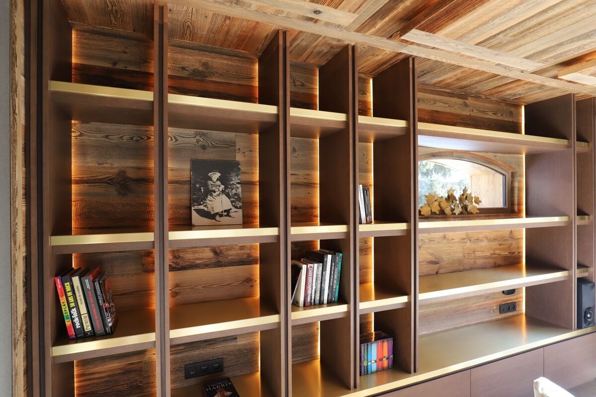 Elegant built-in bookshelf with rustic wood, brass accents, and integrated warm lighting. Books and decor adorn its shelves, matching the wooden ceiling.