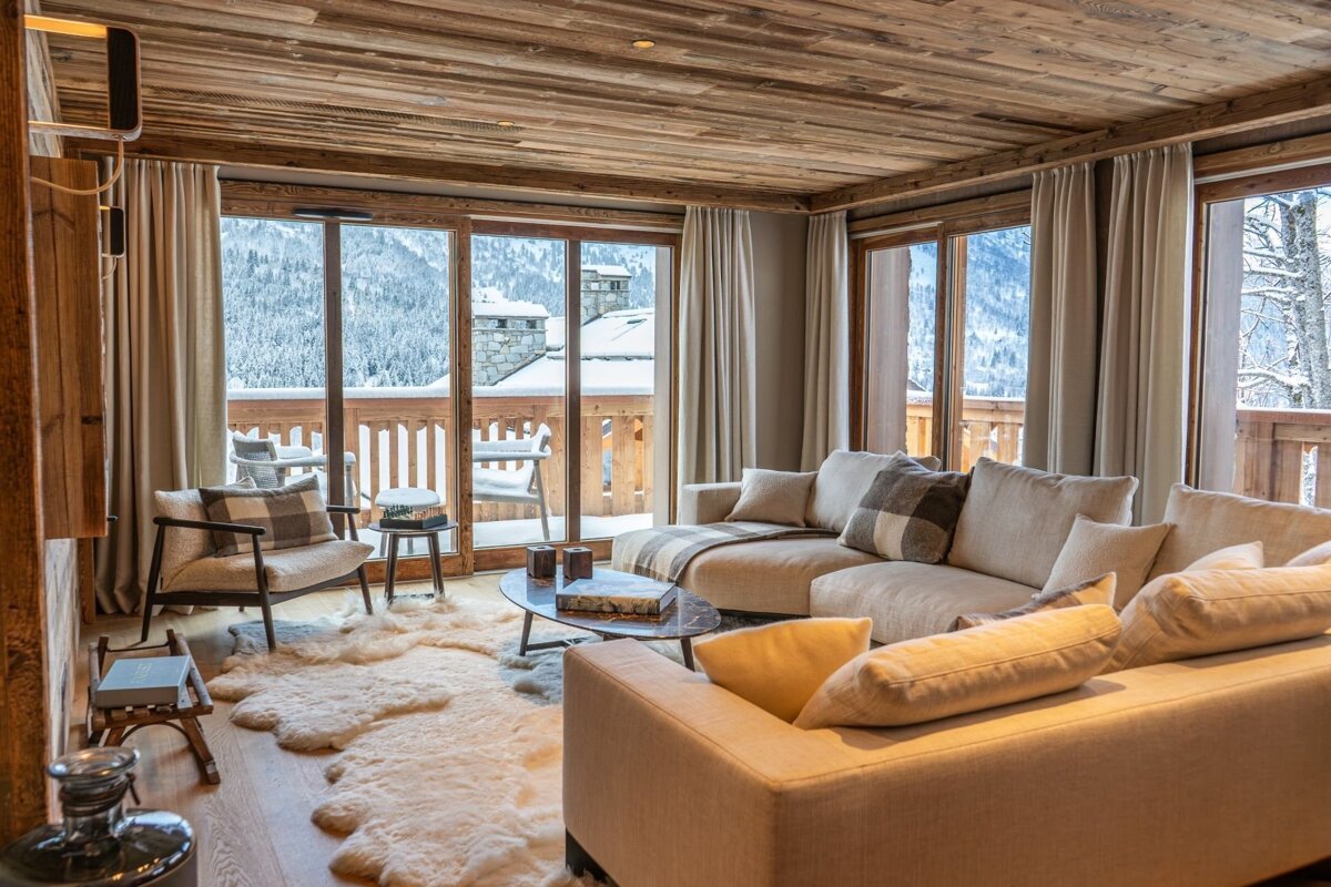 Cozy wooden chalet living room with plush sofas, a sheepskin rug, and large windows offering panoramic views of snow-covered mountains.