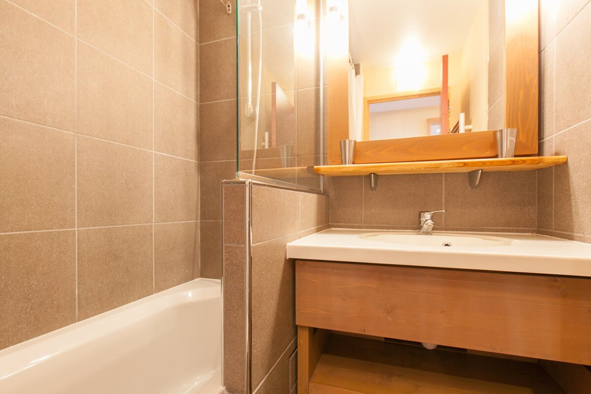 A modern bathroom with a clear glass vessel sink on a wooden counter, a framed mirror, and a shower cabin with dark grey tiles.