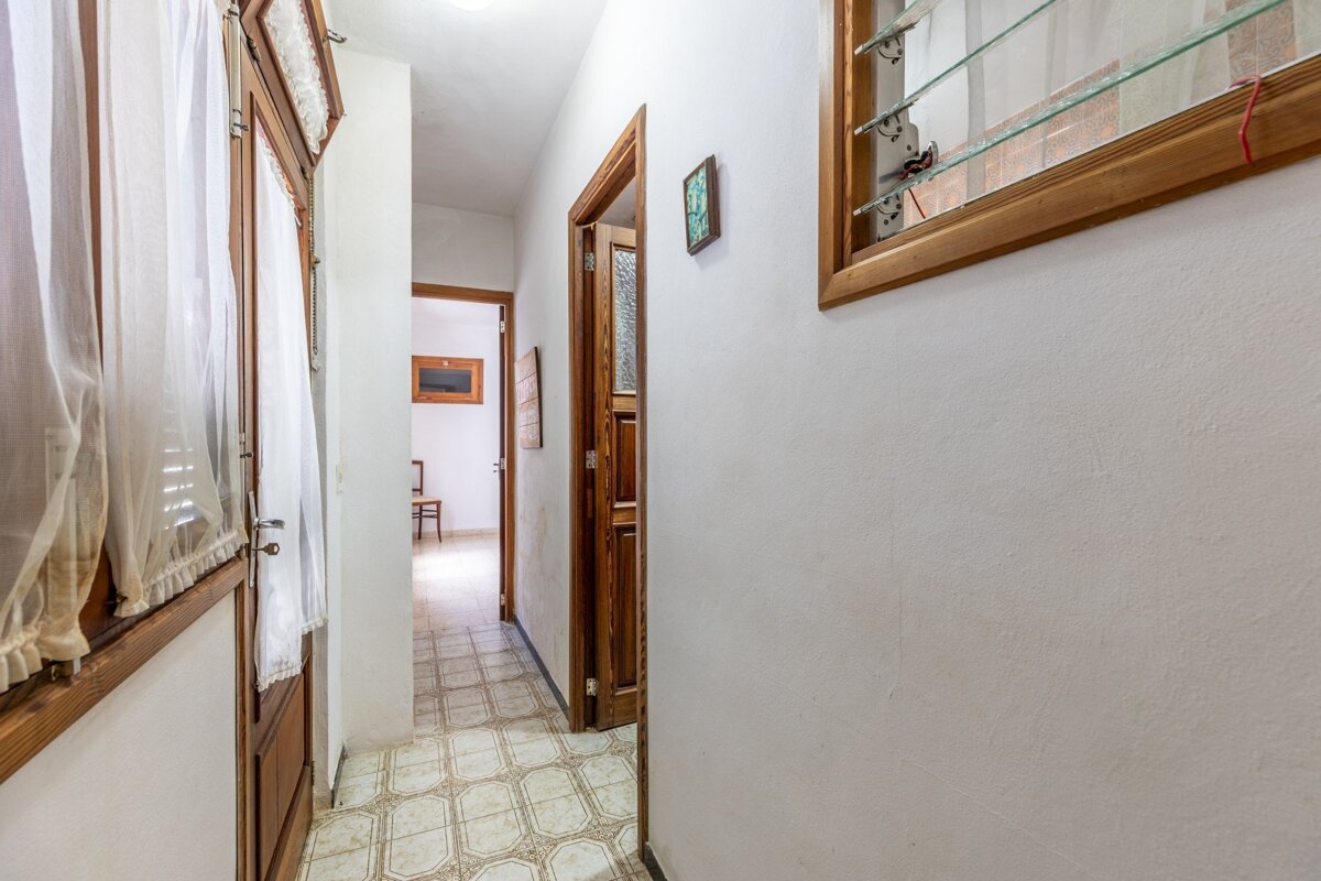 A narrow hallway with white walls and patterned tiled floor. Features wooden doors, a glass-slatted window, and sheer curtains.