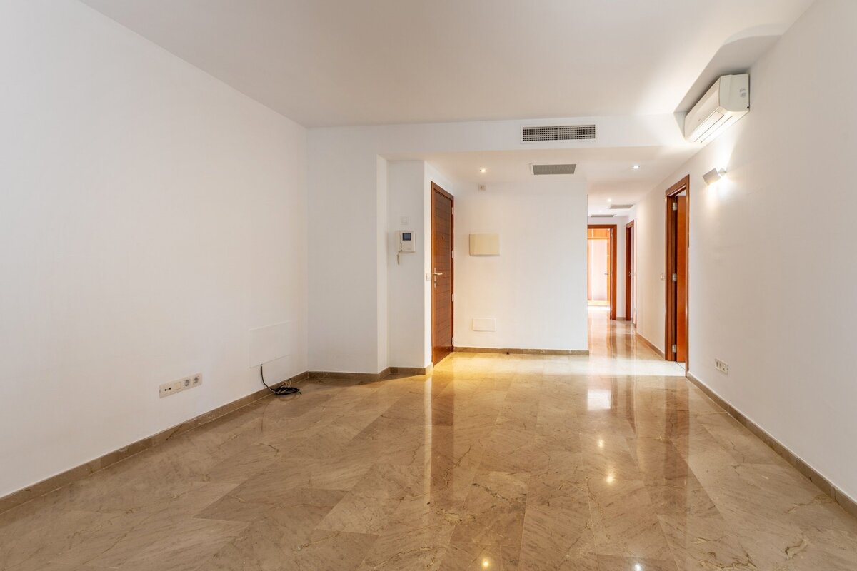 An empty, modern room with white walls, shiny beige marble floors, and dark wooden doors leading to a hallway. Features an AC unit.