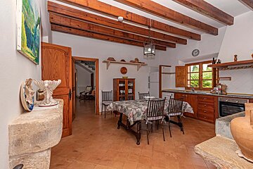 A rustic kitchen-diner with exposed wooden beams, terracotta floor tiles, a dining table, traditional wooden cabinetry, and a view into another room.