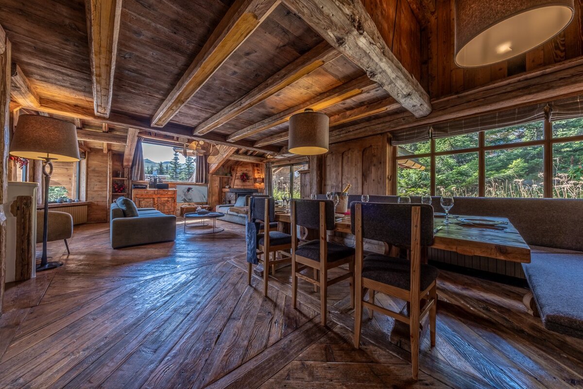 A rustic wooden chalet interior featuring a dining area, living room, and large windows revealing a scenic forest landscape.
