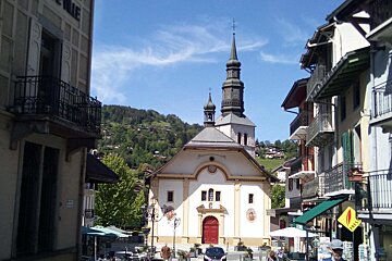 A narrow European street leading to a white church with a tall dark spire, set against a backdrop of green, forested hills.