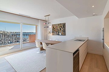 A modern open-plan kitchen and dining area with white decor and a large balcony overlooking a bustling marina filled with boats under a blue sky.