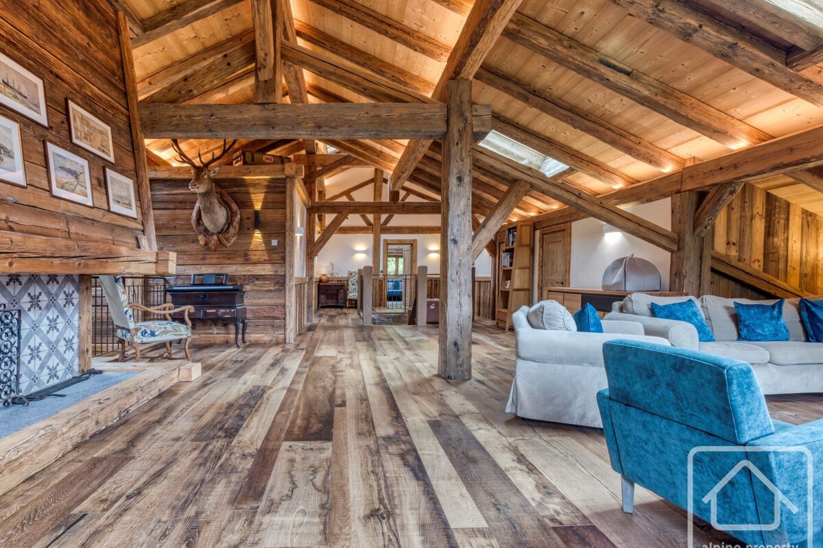 Spacious rustic chalet living room with exposed wooden beams, walls, and floor. Features a fireplace, deer head, and blue-accented sofas.
