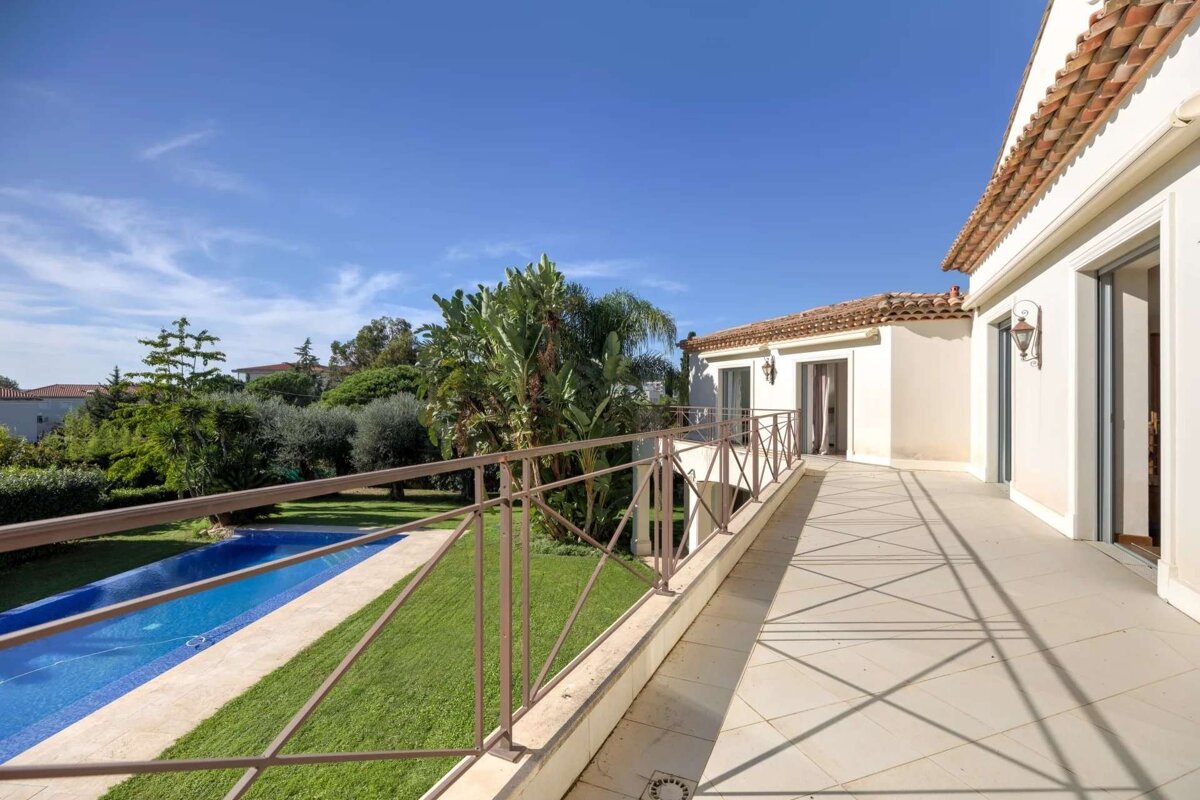 Sunny house balcony with terracotta roof overlooking a long blue pool, green lawn, and lush trees under a clear blue sky.