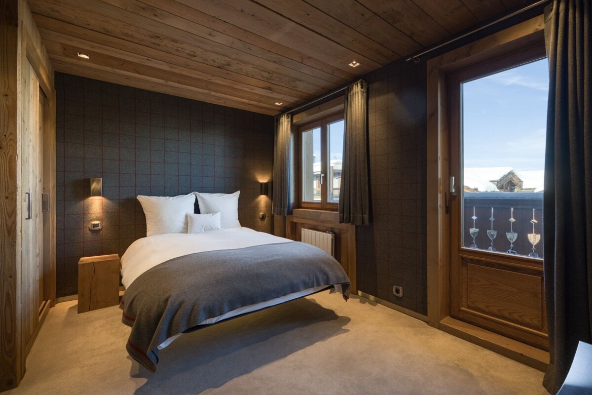 Cozy chalet bedroom with a wooden ceiling, dark patterned wall, white bed, and a balcony door revealing a bright mountain view.