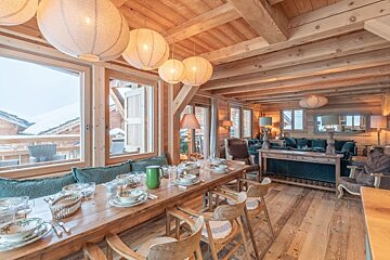 Cozy wooden chalet interior featuring a long set dining table, paper lantern lights, and a snowy mountain view through large windows.