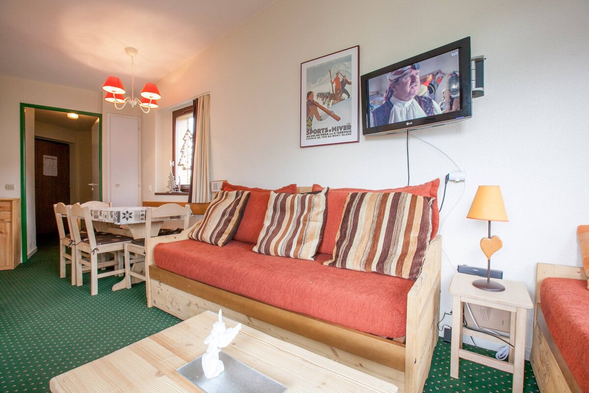 A rustic living/dining room featuring an orange sofa, striped pillows, wooden furniture, green carpet, a TV, and a vintage