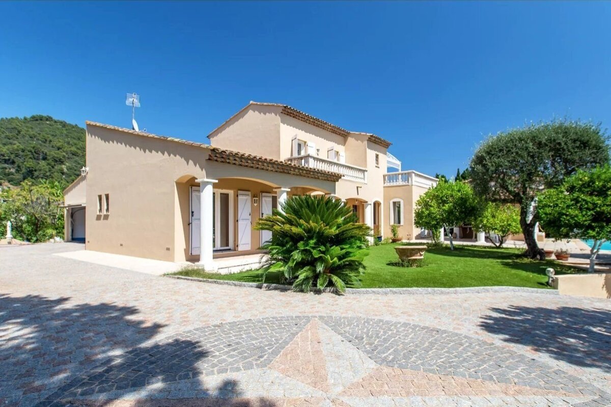 A luxury Mediterranean villa with a paved star-pattern driveway, green lawn, and trees under a clear blue sky.