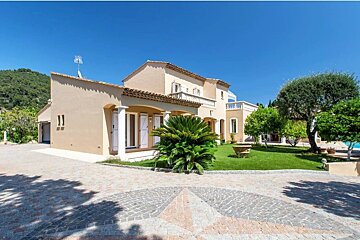A luxury Mediterranean villa with a paved star-pattern driveway, green lawn, and trees under a clear blue sky.
