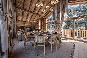 A cozy wooden chalet features a stylish dining table, inviting living space, and stunning window views of an alpine village and trees.