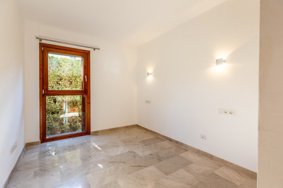 An empty room with white walls, a large wooden-framed window showing greenery, gray marble tile floor, and illuminated wall sconces.
