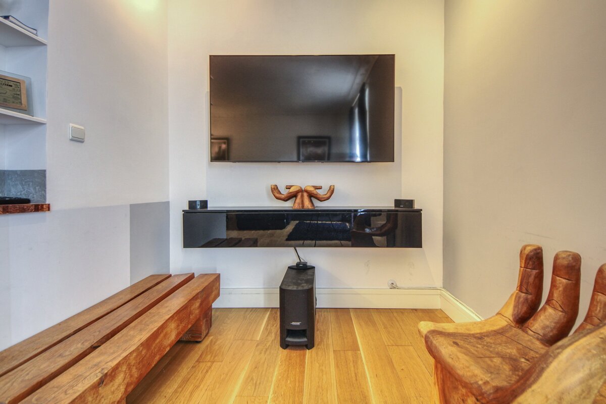 A modern living room with a wall-mounted TV, black floating console, Bose speaker, and rustic wooden seating on a light wood floor.