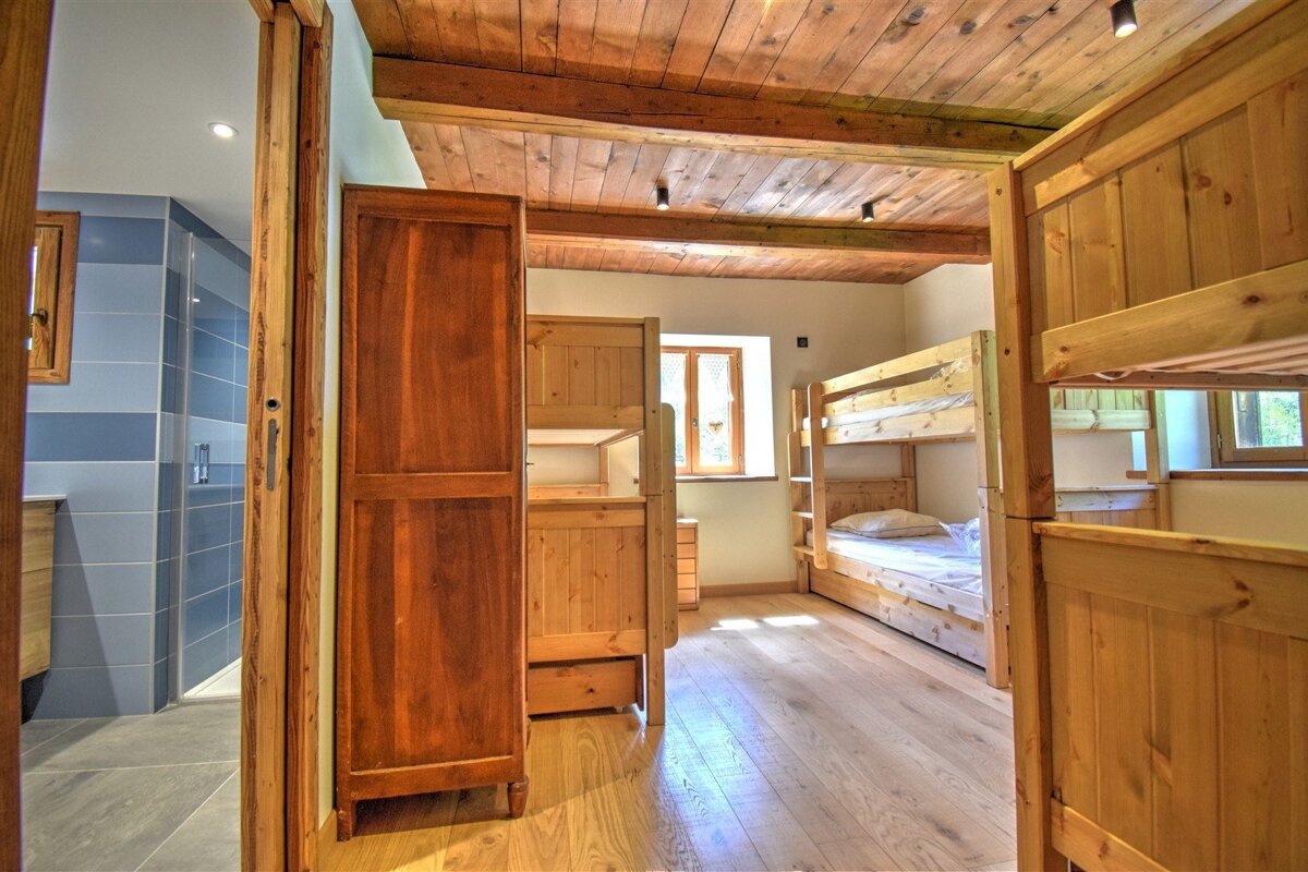 A rustic room with wooden bunk beds, floor, and beamed ceiling, next to a blue-tiled bathroom. Features natural wood furnishings.