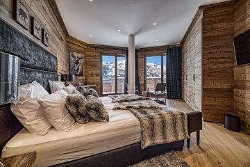 A luxurious, wood-paneled ski chalet bedroom with a large bed, faux fur accents, and stunning panoramic mountain views from large windows.