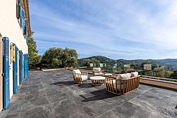 Spacious tiled patio with modern outdoor furniture and a house with blue shutters, boasting panoramic hill views under a clear sky.
