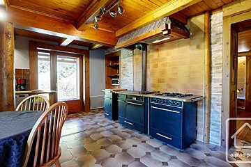 A rustic kitchen with exposed wooden beams, a tiled floor, and a large dark blue range stove. A window offers a view of the outdoors.