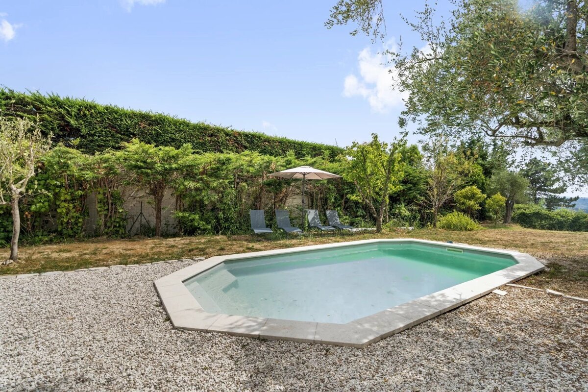 An outdoor swimming pool surrounded by gravel, green hedges, trees, and lounge chairs under an umbrella.