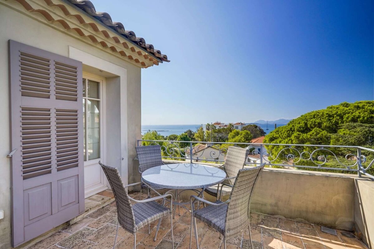 Sunny balcony with outdoor seating, boasting a stunning sea view over green trees, houses, and distant hills under a clear blue sky.