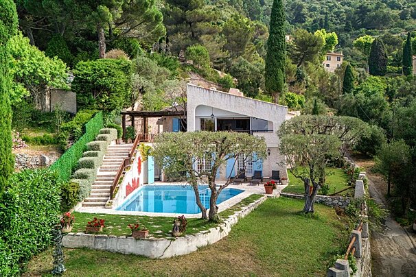 A modern villa with a swimming pool and blue shutters, built into a lush green hillside with terraced gardens and tall cypress trees.