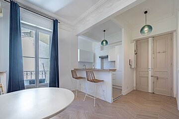 A bright, open-plan apartment interior featuring a dining area, a kitchenette with bar seating, a large window with blue curtains, and light wood floors.
