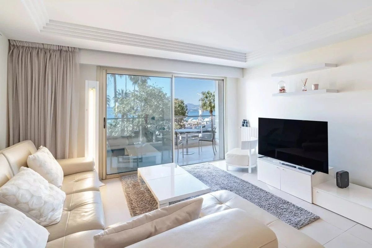 Bright, modern living room featuring white leather sofas, a TV, and sliding glass doors opening to a sunny balcony with palm trees.