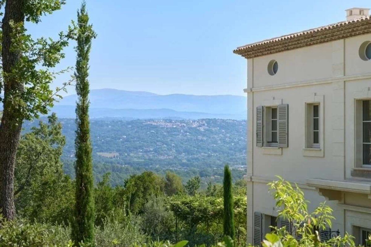A white building with shutters overlooks a lush green valley with a distant town and mountains under a clear blue sky.