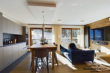 A modern open-plan living space featuring a sleek kitchen, dining table, and blue sofa, all overlooking a snowy mountain landscape through large windows.