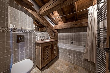 A rustic attic bathroom with exposed wooden beams, grey tiled walls, white toilet, sink, and bathtub. Pebble accents add character to the design.