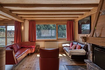 A rustic living room with exposed wood beams, red leather sofas, a fireplace, and large windows looking out to trees.