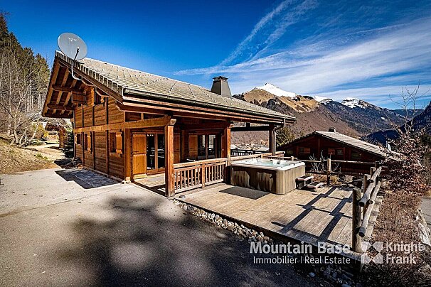 A charming wooden chalet with a hot tub on a deck, offering stunning views of snowy mountains under a bright blue sky.