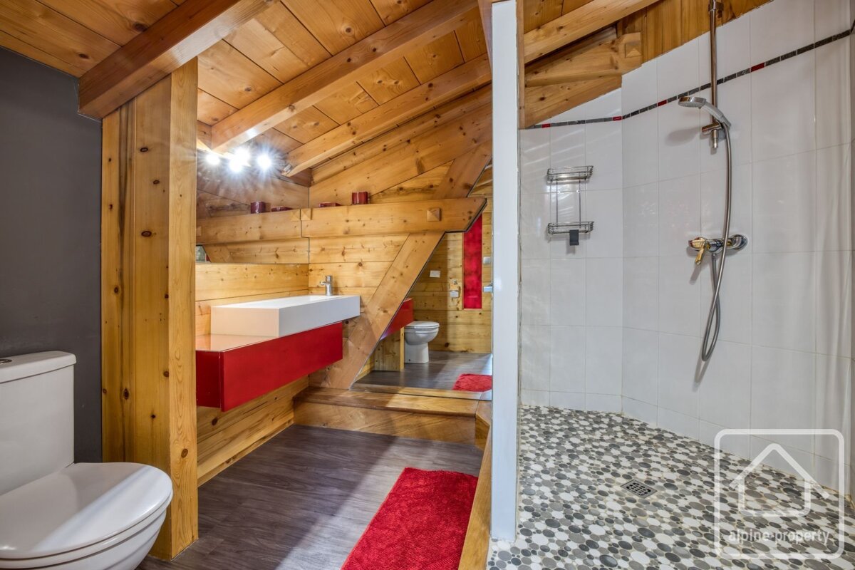 A rustic modern attic bathroom with wood walls, red vanity, white toilet, and a walk-in shower with mosaic tile floor.